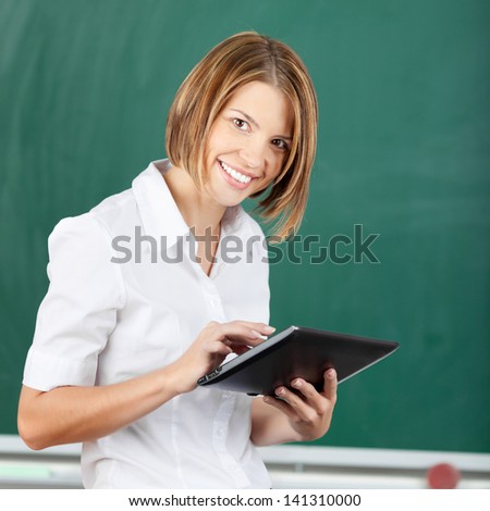 Smiling woman with a tablet-pc standing in front of a school blackboard looking at the camera
