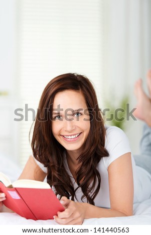 Smiling woman reading in bed lying on her stomach relaxing as she enjoys a lazy day