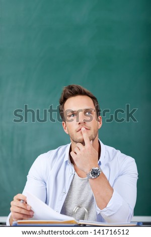 Thoughtful young man sitting in front of chalkboard with finger on chin