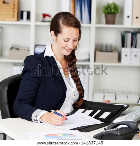 Businesswoman sitting at her desk writing notes in the office with a friendly smile on her face