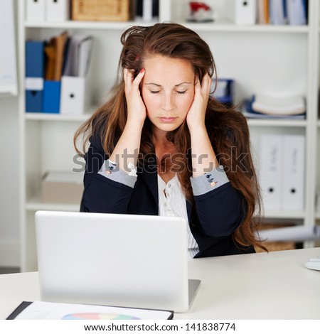 Unhappy woman sitting in her office with her eyes closed and hands to her head either because she is unwell or because she is under stress and frustrated in her job