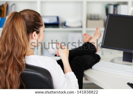 Office worker taking a coffee break with her bare feet raised up on the desk and a cup of espresso in her hands, over the shoulder rear view