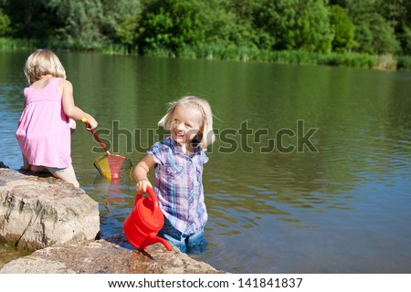 Two cute little blond girls having fun at the lake paddling in the calm water with a fishing net and colourful red plastic watering can