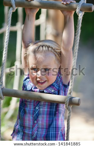 Smiling little girl playing on a rope ladder hanging by her hands from a wooden rung in an outdoor playground