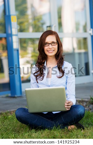 Young woman or student sitting cross legged working on her laptop outdoors on a green lawn in front of a building