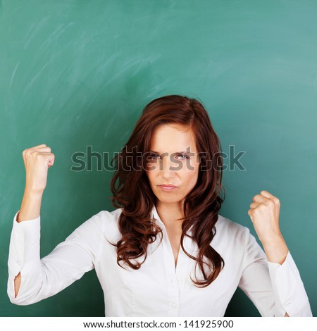 Angry attractive young woman standing against a blank blackboard shaking her fists in the air