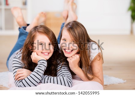 Two happy young best friend teenage girls, smile to the camera, while laying on the bedroom floor over pillows
