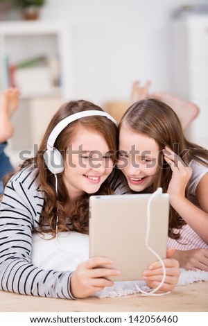 Close-up of two happy young teenage girls sharing headphones connected to a tablet to listen to music while laying on the living-room floor