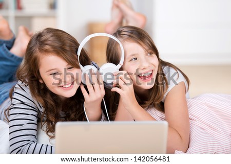 Close-up of two happy young teenage girls sharing headphones connected to a laptop to listen to music while laying on the living-room floor