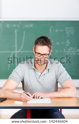 Male thoughtful teacher checking examination papers at bench in classroom