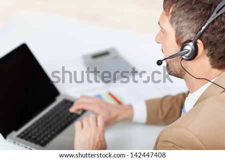 Rear view of mature male customer service executive conversing on headset at desk in office