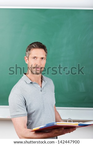Thoughtful mature male teacher holding binder against blank blackboard