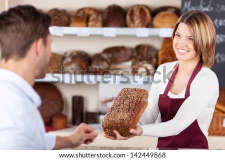 Smiling friendly young bakery assistant selling bread showing a wholewheat loaf to a male customer