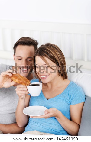 Attractive happy young couple sitting up against the pillows enjoying a delicious coffee and croissant in bed