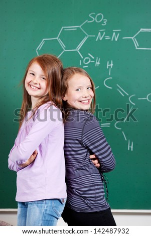 Laughing little girls having fun in school standing back to back in front of the blackboard looking at the camera with lovely smiles