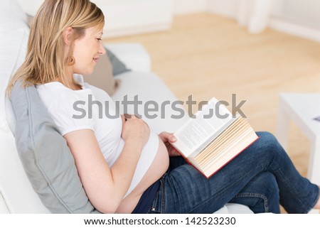Side view of young pregnant woman reading story book while sitting on sofa at home