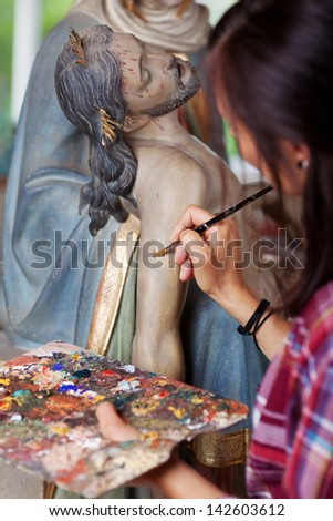 Woman painting Jesus Christ\'s statue with paintbrush in workshop