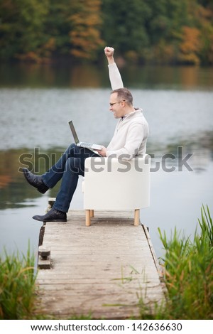 Full side view of excited mature man celebrating victory while using laptop on pier