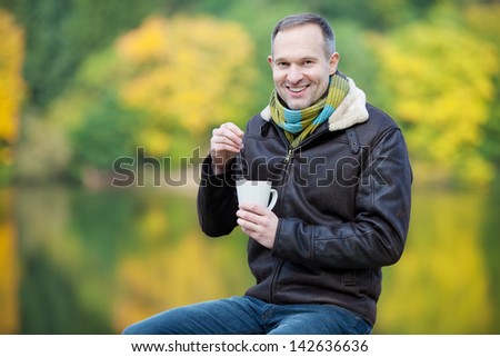 Happy mature man dipping tea bag in cup while sitting on fence against lake