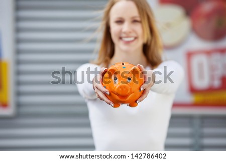 Image of a beautiful young woman showing her piggy bank standing in front of the supermarket.