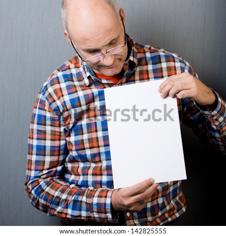 Mature balding man wearing casual clothing holds a blank white paper sheet with copy-space in front of the chest and looks down at it, with a gray wall as background
