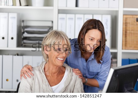 Successful team at the office consisting of an elderly and young woman standing arm in arm studying a desktop monitor with smiles of anticipation