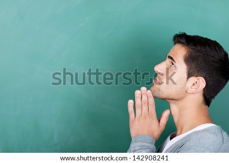 Closeup of young male student with hands clasped looking away against chalkboard in classroom