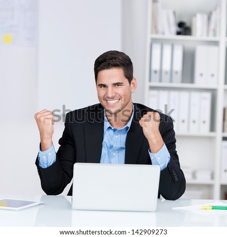 Portrait of happy young businessman with laptop celebrating victory at desk in office