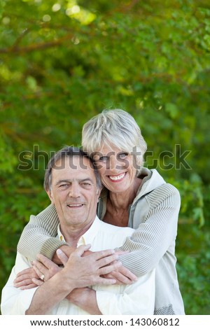 Senior woman embracing her husband at the back over the green trees background