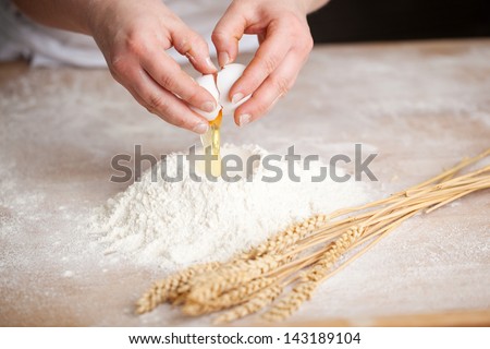 Hands of a female bakery chef breaking an egg into a heap of measured flour while making bread with a bunch of wheat alongside