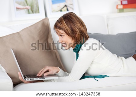 Smiling woman browsing the internet on her laptop computer while lying on her stomach on a sofa