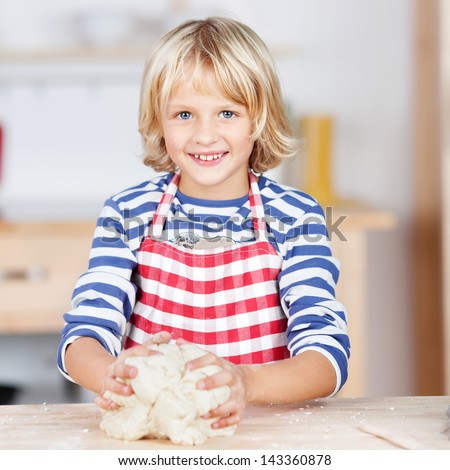 Happy little blond girl in a red and white striped apron kneading dough at a kitchen counter while baking biscuits and cookies