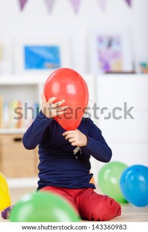 Playful little girl kneeling on the floor hiding her face behind a red balloon