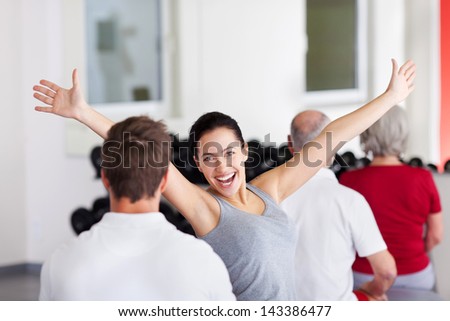 Happy excited young woman with arms raised sitting with group in gym