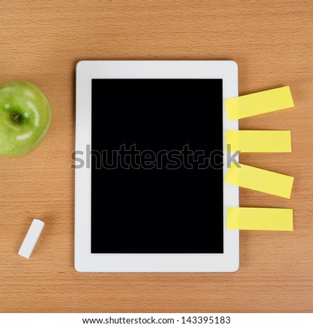 Digital tablet with a black empty display with post-it notes stuck on the side over a school desk next to an eraser and a green apple