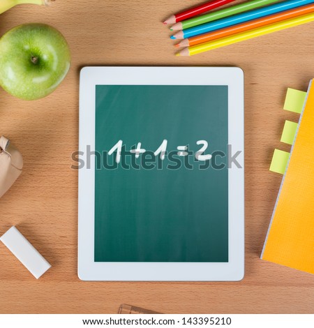 Digital tablet on a school desk with math exercise between a paper notebook, pencils, an eraser, and an apple