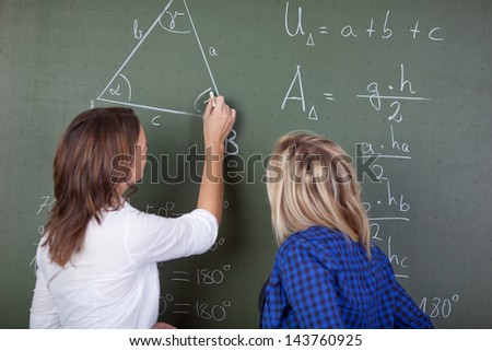 Female teacher solving a mathematics question on the blackboard while a female student standing behind and looking.