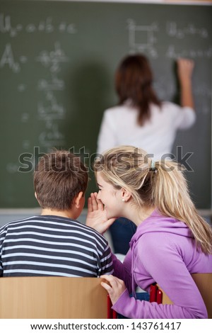 Female student whispering in friend\'s ear while teacher writing on blackboard at classroom