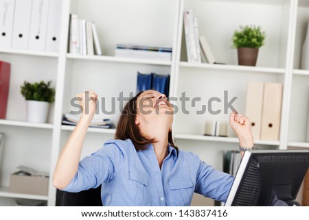 Businesswoman rejoicing in her office sitting at her desk with her head tilted back and a big smile of jubilation and her clenched fists raised in the air