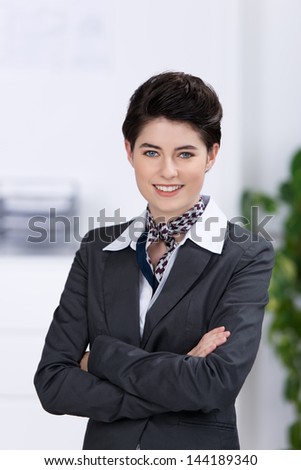 Confident beautiful young businesswoman in a stylish suit and necktie standing with folded arms in the office