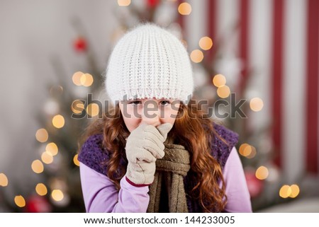 Young girl with a Christmas cold and flu standing in front of the Christmas tree in a warm knitted cap, scarf and gloves coughing
