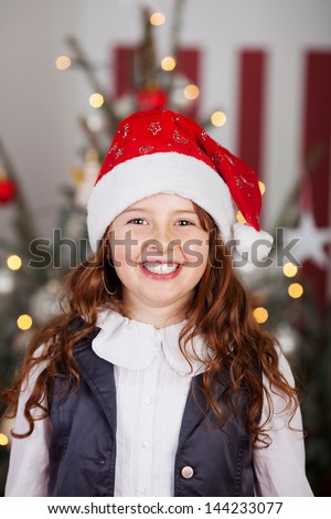 Excited young girl in a red Santa hat standing in front of a Christmas tree decorated with twinkling lights in the living room
