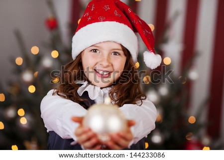 Beautiful young girl in a colorful red Santa hat holding out a white Christmas bauble in her outstretched hands, focus to her face with decorated tree backdrop