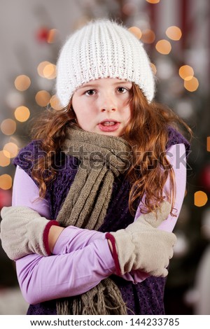 Shivering cold young girl standing in front of a decorated Christmas tree in a warm knitted woolly cap and scarf rubbing her arms with her gloved hands