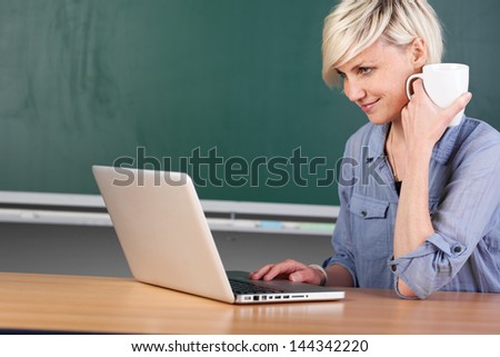 Young female teacher with laptop and coffee cup sitting at school table