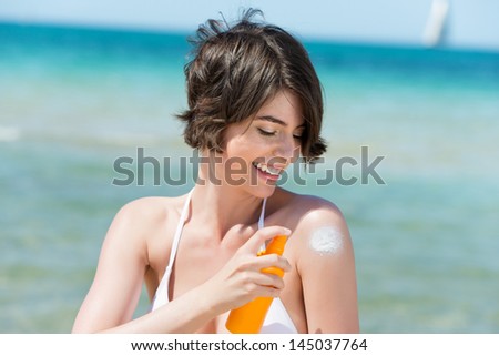 Laughing woman applying suntan lotion to her shoulder from a spray bottle as she stands on the seashore overlooking the ocean