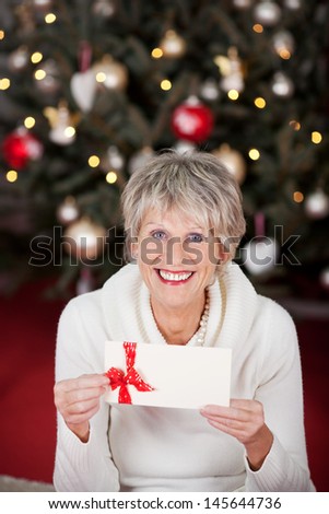 Smiling beautiful senior lady with a gift voucher displayed in her hands sitting in front of a decorated Christmas tree with twinkling lights