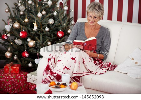 Elderly attractive woman relaxing under a colorful blanket with reindeer on the sofa reading a book in front of the Christmas tree