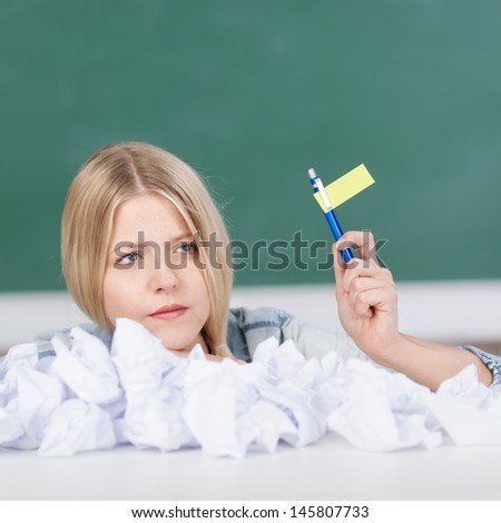 Young woman holding flag made of pen and tag with heap of paperballs on desk in classroom