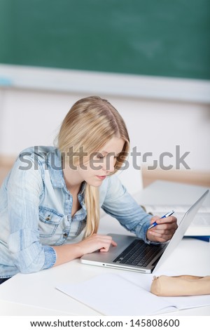 Young blond female student working on a laptop concentrating on reading the screen while holding a pen in her hand ready to take notes